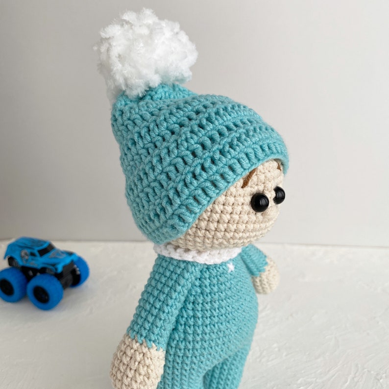 May include: A crocheted doll wearing a blue hat with a white pom-pom. The doll is wearing a blue crocheted outfit and has brown yarn hair.