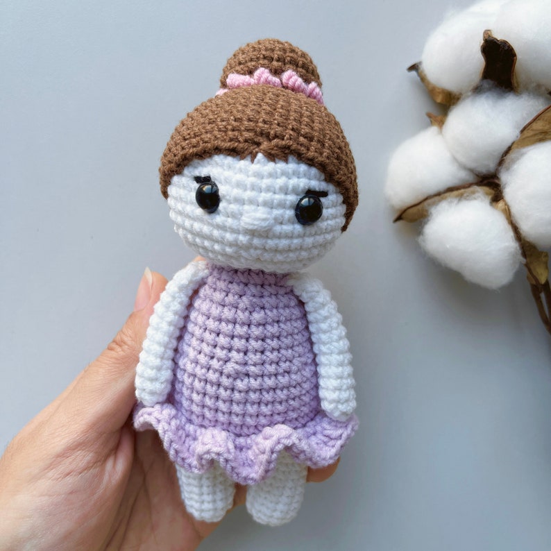 Puede incluir: Una mu&ntilde;eca de crochet con cabello casta&ntilde;o, una diadema rosa y un vestido lavanda. La mu&ntilde;eca tiene una cara blanca con ojos negros y una peque&ntilde;a sonrisa.