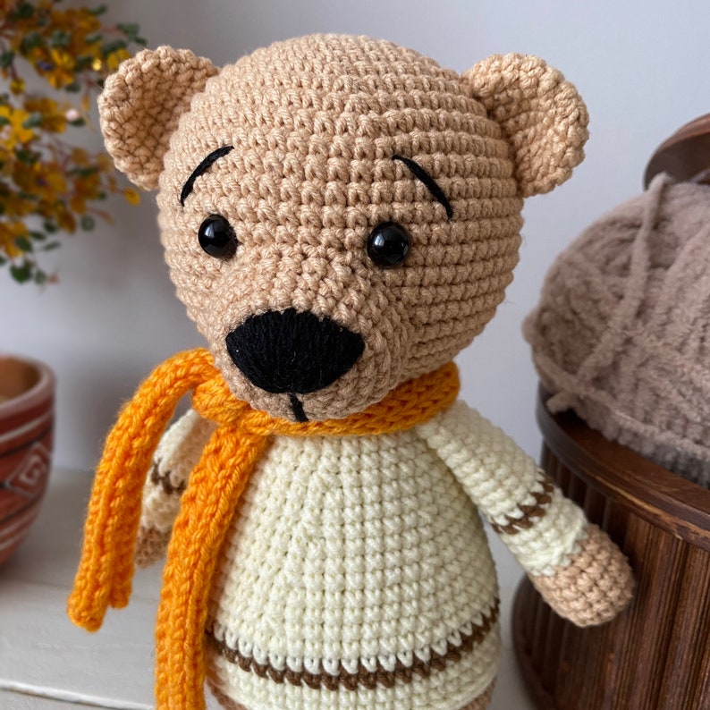 Peut inclure: Un ours en peluche crochet&eacute; portant un pull blanc et une &eacute;charpe orange. L'ours a des yeux bruns et un nez noir.