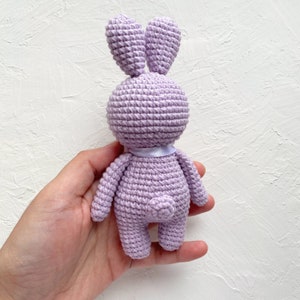 May include: A crocheted lavender bunny rabbit toy with a white ribbon around its neck. The bunny is standing on its hind legs with its front paws tucked in.