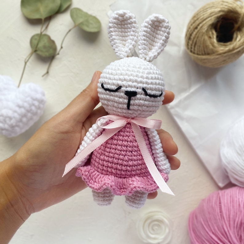 May include: A white crocheted bunny with pink eyes and a pink crocheted dress. The bunny is holding a pink ribbon in its paws.