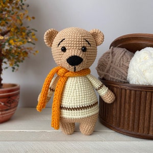 Peut inclure: Un ours en peluche crochet&eacute; portant un pull ray&eacute; marron et blanc et une &eacute;charpe orange. L'ours a des yeux noirs et un nez noir.