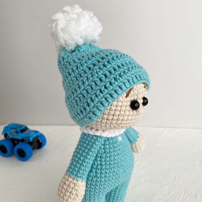 K&ouml;nnte beinhalten: Eine geh&auml;kelte Puppe mit einem blauen Hut mit wei&szlig;em Bommel. Die Puppe tr&auml;gt ein blaues H&auml;kelkleid und hat braunes Garnhaar.
