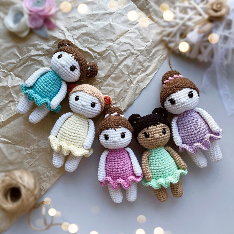 Puede incluir: Cinco mu&ntilde;ecas de crochet con vestidos y cabello de diferentes colores. Las mu&ntilde;ecas est&aacute;n dispuestas en c&iacute;rculo sobre una superficie blanca.