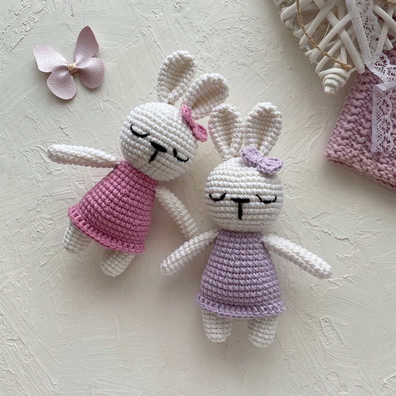 May include: Two crocheted white bunny plushes with pink and purple dresses. The bunnies have black eyes and noses and are lying on a white surface.