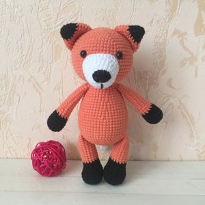 May include: A crocheted orange fox plush toy with black ears, paws, and nose. The toy has white eyes and a white muzzle.