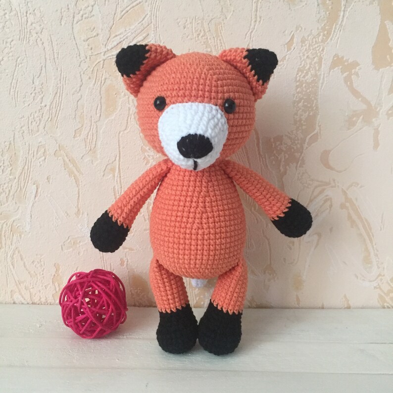 May include: A crocheted orange fox plush toy with black ears, paws, and nose. The toy has white eyes and a white muzzle.
