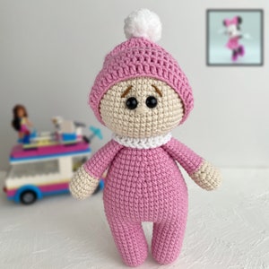 May include: A pink crocheted doll wearing a pink and white hat with a white pom-pom. The doll has brown embroidered eyes and a white crocheted collar.