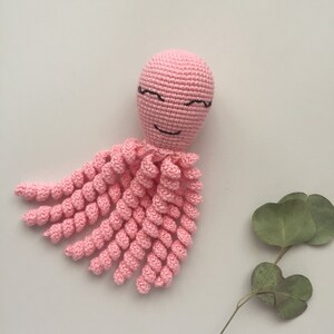 May include: A pink crocheted octopus with a smiling face and black eyes. The octopus has eight tentacles that are curled.