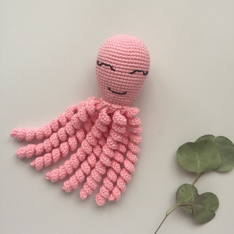 May include: A pink crocheted octopus with a smiling face and black eyes. The octopus has eight tentacles that are curled.