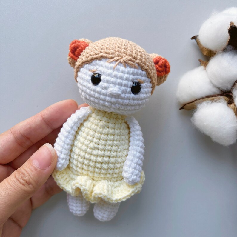 Puede incluir: Una mu&ntilde;eca de crochet con un vestido amarillo y lazos de pelo naranja. La mu&ntilde;eca tiene ojos negros y una cara blanca.