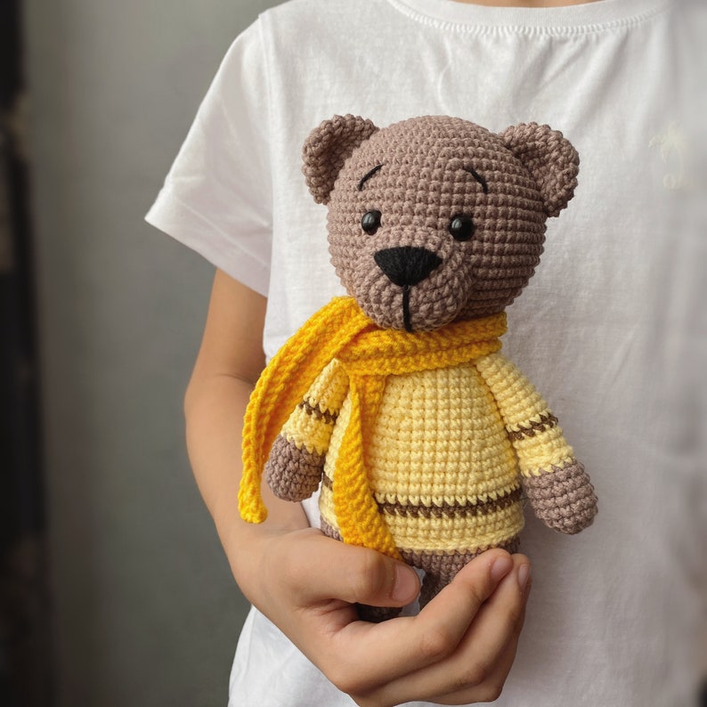 Peut inclure: Un ours en crochet marron portant une &eacute;charpe jaune. L'ours tient une &eacute;charpe jaune dans ses pattes.
