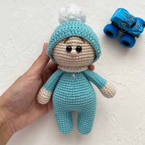 May include: A crocheted baby doll wearing a blue hat with a white pom-pom. The doll has brown eyes and is wearing a blue outfit.