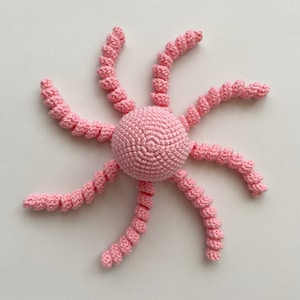 May include: A pink crocheted octopus with eight curled tentacles. The octopus is made of soft yarn and is perfect for a baby's toy.