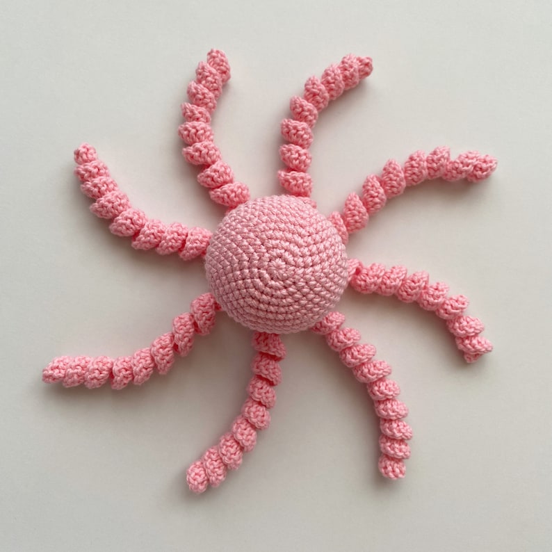 May include: A pink crocheted octopus with eight curled tentacles. The octopus is made of soft yarn and is perfect for a baby's toy.