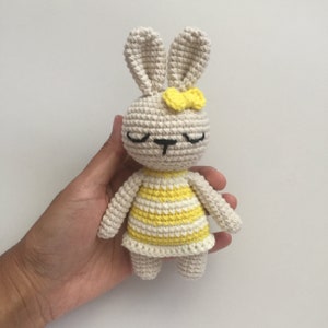 May include: A crocheted stuffed bunny toy with a yellow and white striped dress and a yellow bow. The bunny has closed eyes and a black nose.