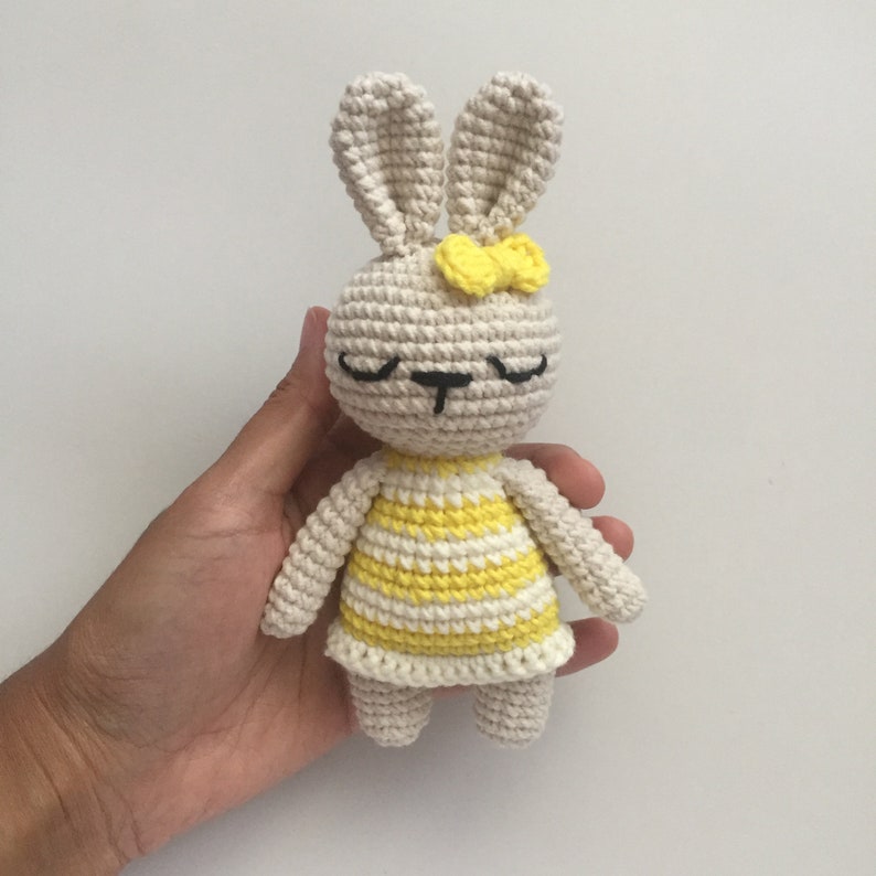 May include: A crocheted stuffed bunny toy with a yellow and white striped dress and a yellow bow. The bunny has closed eyes and a black nose.