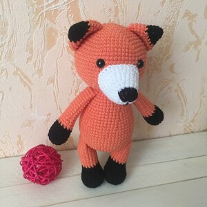 May include: A crocheted orange fox plush toy with black ears, black feet, and a white face with a black nose. The toy is sitting on a white surface.