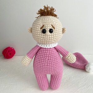 May include: A crocheted baby doll with brown hair, wearing a pink outfit and a white collar. The doll has black eyes and a sad expression. A pink crocheted hat is lying next to the doll.