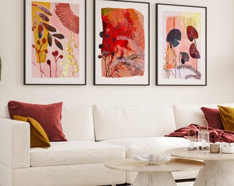 May include: A set of three framed wall art prints featuring abstract botanical illustrations in shades of red, pink, yellow, and dark green. The prints are displayed in thin black frames above a white sectional sofa with red and yellow pillows.