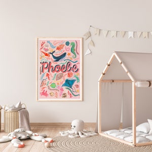 May include: A rectangular framed wall art print with a light pink background featuring the name Phoebe in large pink letters. The design includes colorful illustrations of a whale, seahorse, stingray, crab, fish, shells, and seaweed.