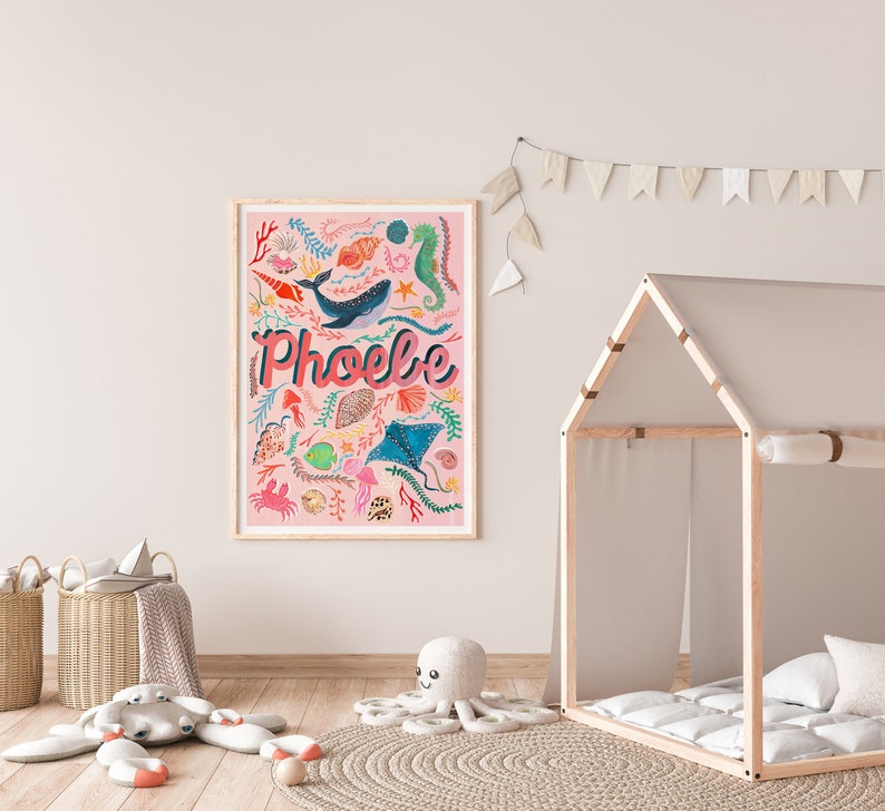 May include: A rectangular framed wall art print with a light pink background featuring the name Phoebe in large pink letters. The design includes colorful illustrations of a whale, seahorse, stingray, crab, fish, shells, and seaweed.