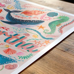 May include: A rectangular art print on a light pink background featuring a dark blue whale, a green seahorse, various seashells, and coral illustrations. The partial text "tha" is printed in large, bold, pink and teal letters across the lower section.