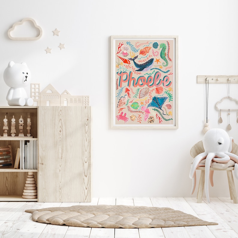 May include: A rectangular wall print in a light wood frame featuring the name Phoebe in pink and blue letters, surrounded by colorful illustrations of a whale, seahorse, stingray, crab, various seashells, and seaweed on a light beige background.