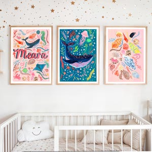 May include: Three framed wall art prints featuring marine life. The left print has a pink background with the name Meara, a whale, and seashells. The center print shows a blue background with a large whale and jellyfish. The right print displays seashells.