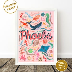 May include: A framed print in a white wood grain frame featuring a pink background with the name Phoebe in large letters. The artwork displays various colorful marine life including a whale, seahorse, stingray, crab, jellyfish, and seashells.