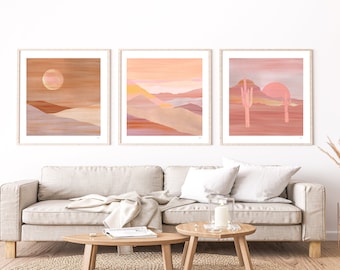 Op de afbeelding: Drie ingelijste abstracte landschapsdrukken met een woestijnthema. De prints hebben een roze en oranje kleurenpalet met geometrische vormen en lijnen. De prints hangen aan een witte muur boven een beige bank.