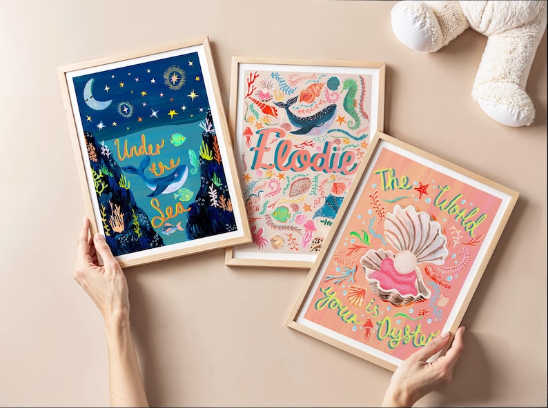May include: Three framed art prints featuring marine themes. One print shows a whale in a dark blue ocean with the text Under the Sea. Another displays the name Elodie with sea creatures. The third shows an open oyster with the text The World is your Oyster.