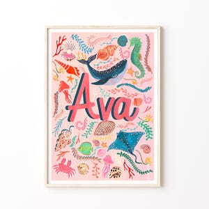 May include: A rectangular wall art print featuring the name Ava in large pink letters centered on a light pink background. The design includes colorful illustrations of a blue whale, a stingray, a green seahorse, a pink crab, and various shells.