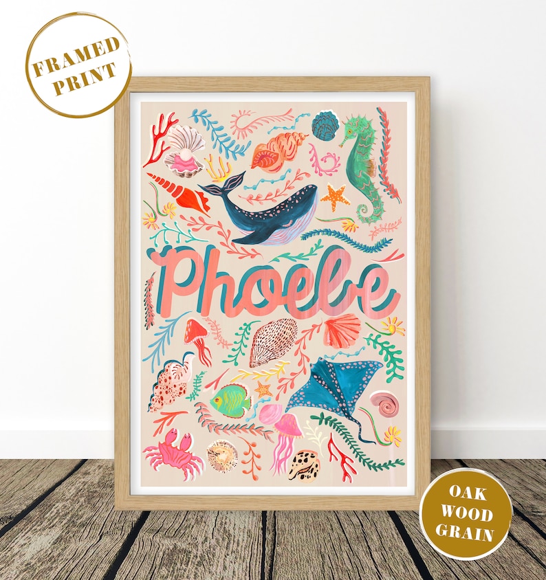 May include: A framed print in an oak wood grain frame featuring a light beige background with a colorful illustration of a whale, seahorse, stingray, crab, fish, jellyfish, and various seashells and coral, with the name Phoebe in pink text.