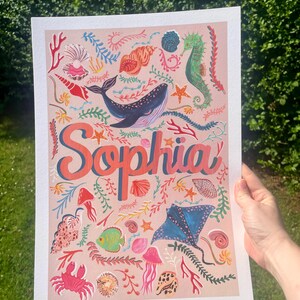 May include: A rectangular art print on a light pink background featuring the name Sophia in large coral-colored letters. The design includes various marine life illustrations such as a blue whale, a green seahorse, a blue stingray, a red crab, jellyfish, and assorted seashells and coral in vibrant colors.