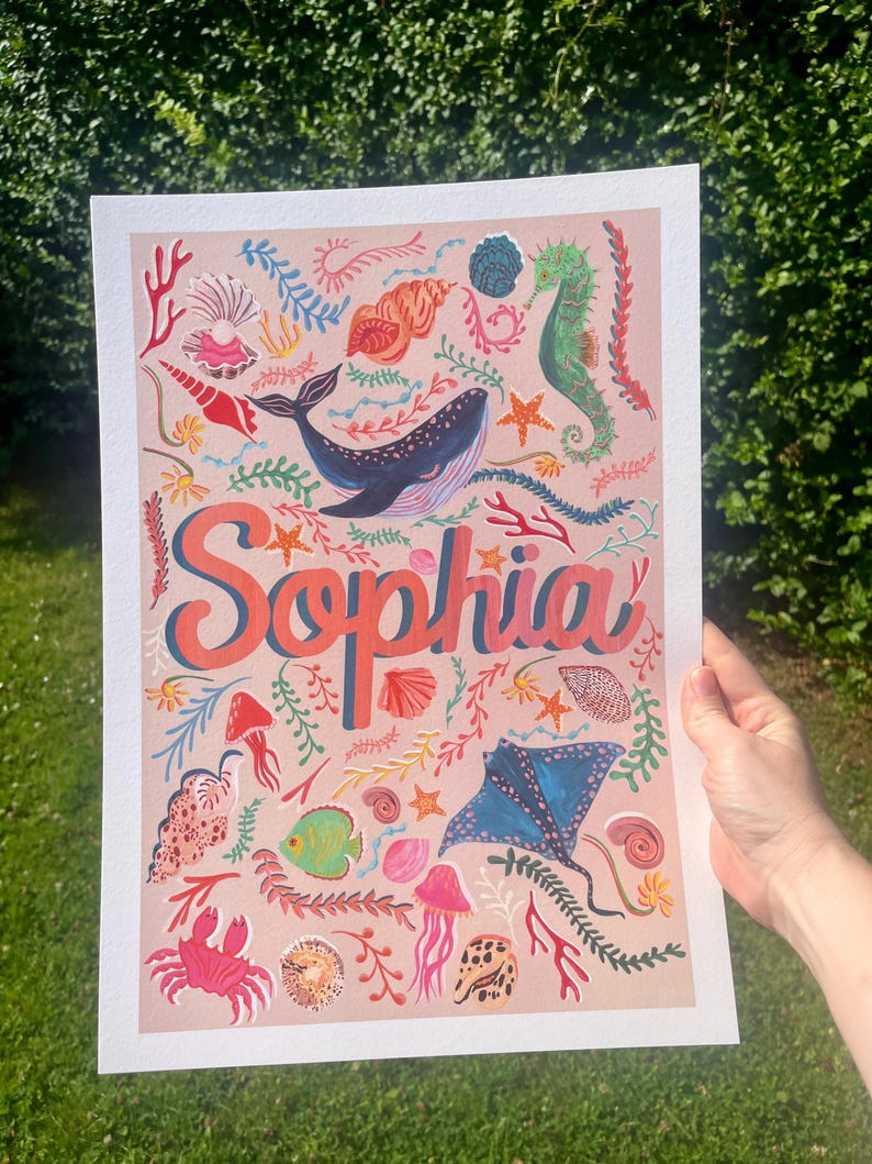 May include: A rectangular art print on a light pink background featuring the name Sophia in large coral-colored letters. The design includes various marine life illustrations such as a blue whale, a green seahorse, a blue stingray, a red crab, jellyfish, and assorted seashells and coral in vibrant colors.