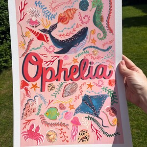 May include: A rectangular art print on a light pink background featuring the name Ophelia in large red letters. The design includes illustrations of a blue whale, a green seahorse, a blue stingray, a pink crab, jellyfish, and various seashells.