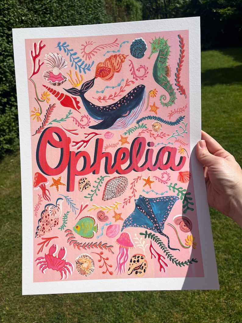 May include: A rectangular art print on a light pink background featuring the name Ophelia in large red letters. The design includes illustrations of a blue whale, a green seahorse, a blue stingray, a pink crab, jellyfish, and various seashells.