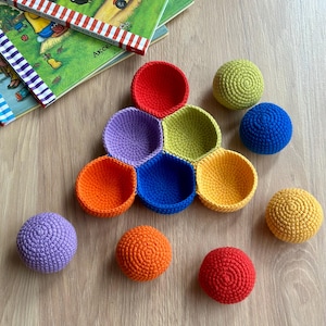 May include: A collection of colorful, crocheted toys, including a set of open-topped bowls and several solid spheres. The bowls are arranged in a pyramid shape, with colors like red, purple, and yellow. The spheres are in matching colors.