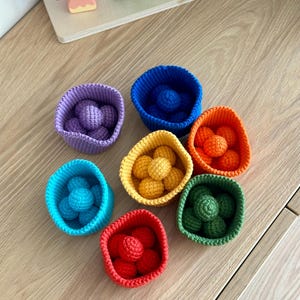 Könnte beinhalten: Eine Sammlung farbenfroher, gehäkelter Schalen, jede gefüllt mit passenden gehäkelten Kugeln. Die Schalen sind in verschiedenen Farben erhältlich, darunter Lila, Blau, Orange, Gelb, Grün, Türkis und Rot. Im Hintergrund ein Holzspielzeug mit Pilz- und Baumformen.