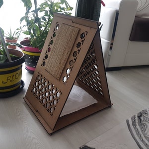 May include: A wooden cat bed shaped like a triangular house with a cutout design. The bed has a soft cushion inside and a scratching post on the side.