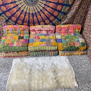 May include: Three colorful patchwork floor cushions with a green border are arranged in a row. The cushions are made of various fabrics in shades of pink, yellow, blue, and green. A white, fluffy rug is in front of the cushions.