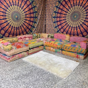 May include: A colorful patchwork sectional sofa with a white rug in front. The sofa is composed of several square cushions in various colors and patterns. The background features a tapestry with a mandala design.