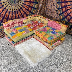 May include: A colorful, patchwork sectional sofa with multiple cushions. The cushions feature a variety of patterns and colors, including pink, yellow, blue, and green. A small, white, fluffy rug sits in front of the sofa. The background includes a tapestry.