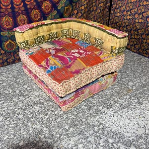 May include: A patchwork floor cushion with a curved backrest. The cushion features a vibrant mix of red, pink, and yellow fabrics. The base is a stack of two cushions, with the top one having a patchwork design and the bottom one a floral pattern.
