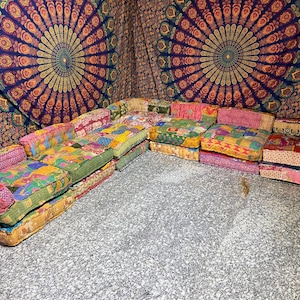 May include: A patchwork sectional sofa with a variety of colorful fabric patterns. The sofa is composed of several individual cushions and armrests, arranged in an L-shape. The fabrics feature floral and geometric designs in shades of pink, yellow, green, and blue.