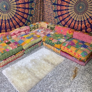 May include: A colorful patchwork sectional sofa with a white fluffy rug in front. The sofa is made of various fabrics in shades of pink, green, yellow, and blue. The background features a tapestry with a mandala design.