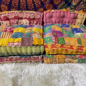 May include: Two square floor cushions with patchwork designs. The cushions feature a variety of colors, including yellow, red, green, and blue. The top of each cushion is a solid color, and the sides have a striped pattern.