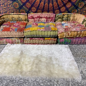 May include: Three floor cushions with patchwork fabric in various colors, including red, yellow, and green. The cushions are arranged in a row, with a white fluffy rug in front.