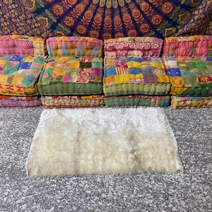 May include: A collection of colorful, patchwork floor cushions with a rectangular, cream-colored rug in front. The cushions are made of various fabrics in shades of pink, green, yellow, and blue. The rug has a soft, fluffy texture.